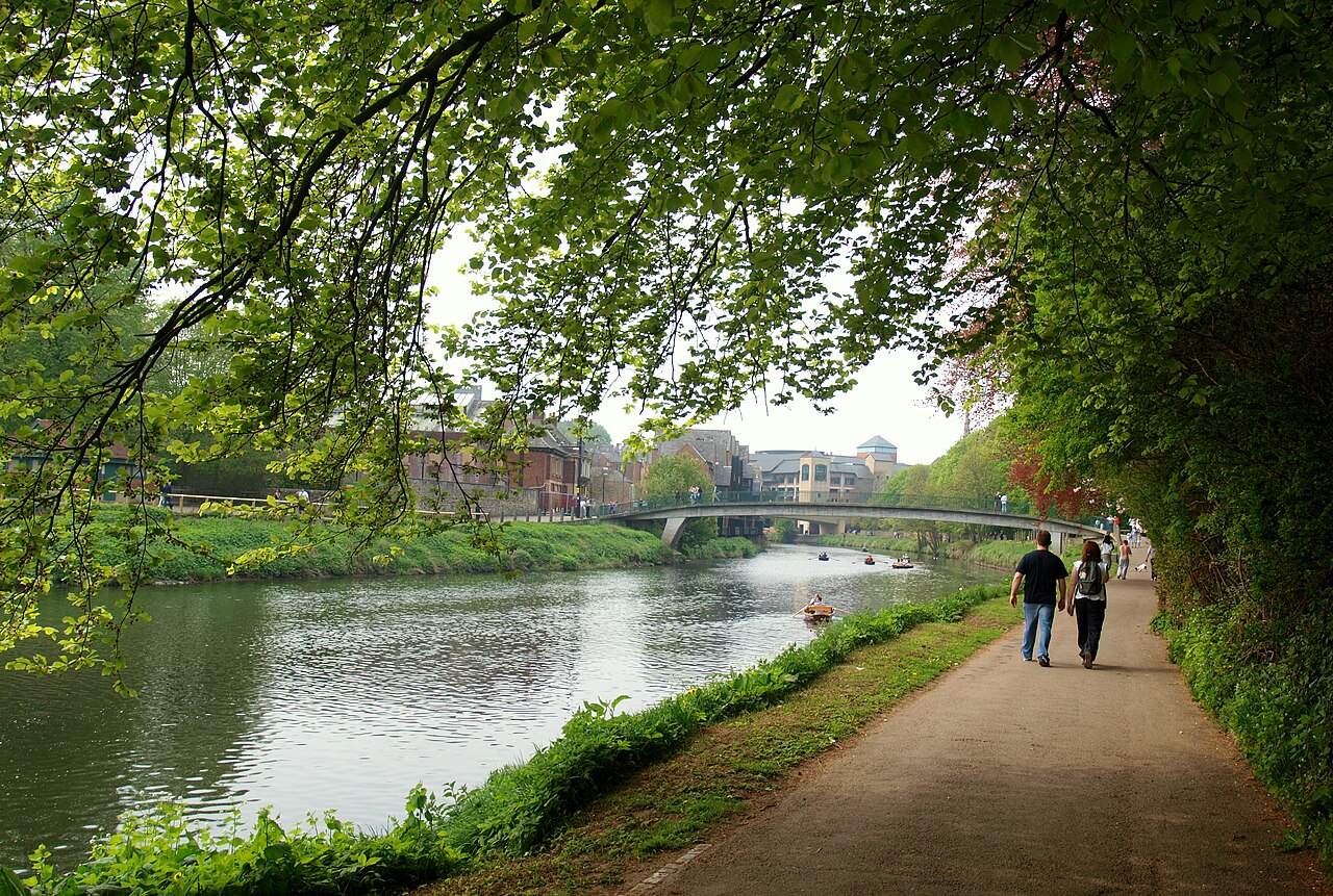 The riverside path along the Wear at Durham, under heavy trees, with a footbridge and the city beyond.
